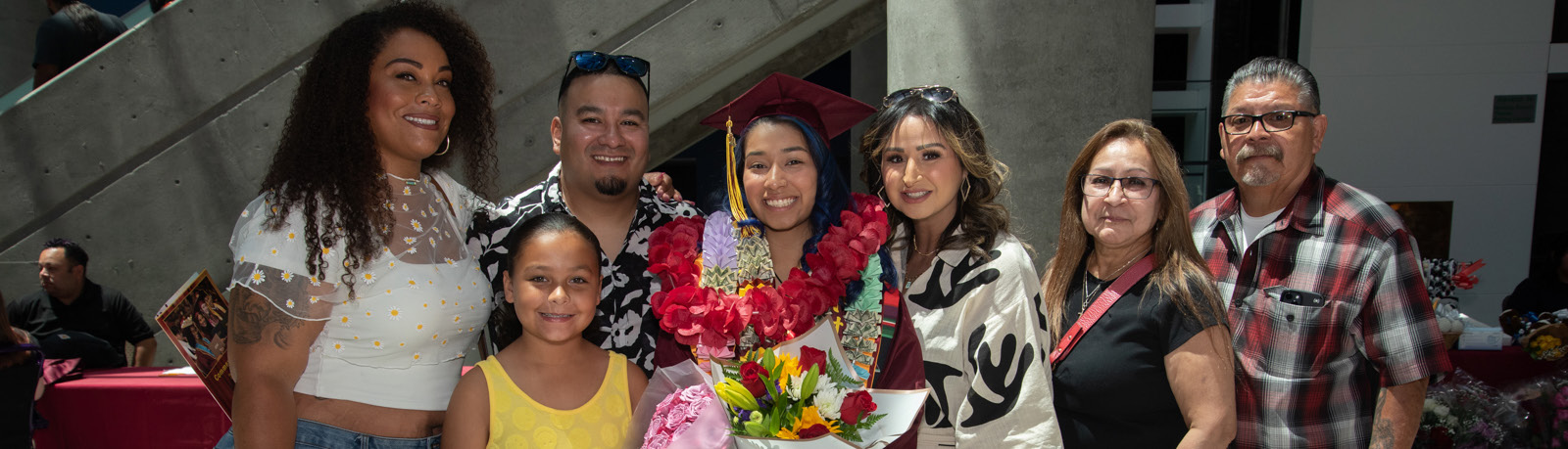 A smiling graduate in a cap and gown, wearing a large flower lei and holding a bouquet of flowers, poses with a group of six family members and friends