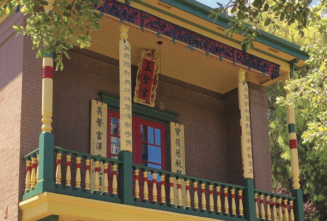 The Ng Shing Gung temple replica today. Photo courtesy of The Californian, Number 43, Oct 2016. A picture of a Chinese-style temple balcony, painted in vibrant red, green, and yellow tones and surrounded by green foliage