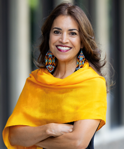 Dr. Rochelle Gutiérrez wearing colorful beading earrings and a yellow scarf, smiling with her arms crossed